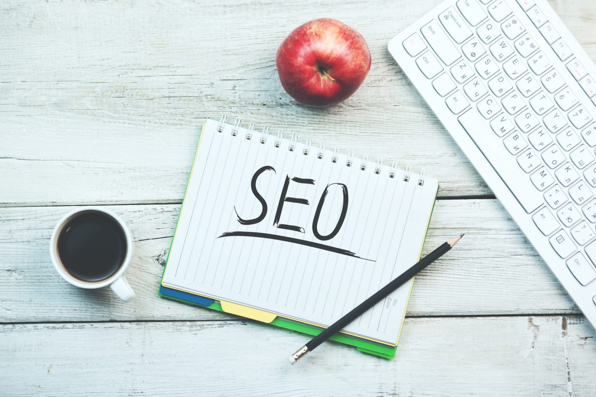 The word SEO is written on a notebook next to an apple and a keyboard, hinting at the importance of SEO strategies for online visibility.