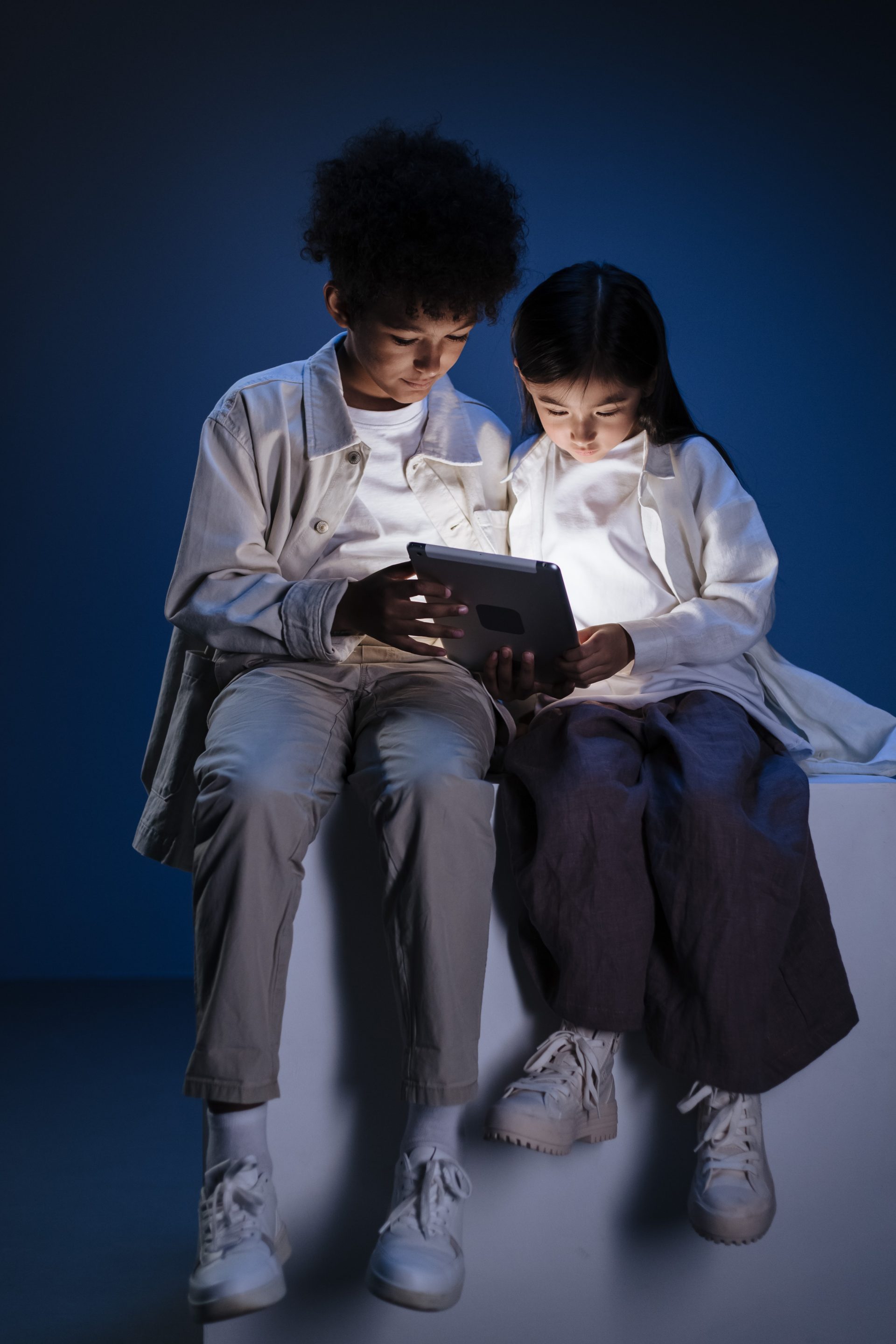 Two children sitting on a white cube using a tablet for social media.