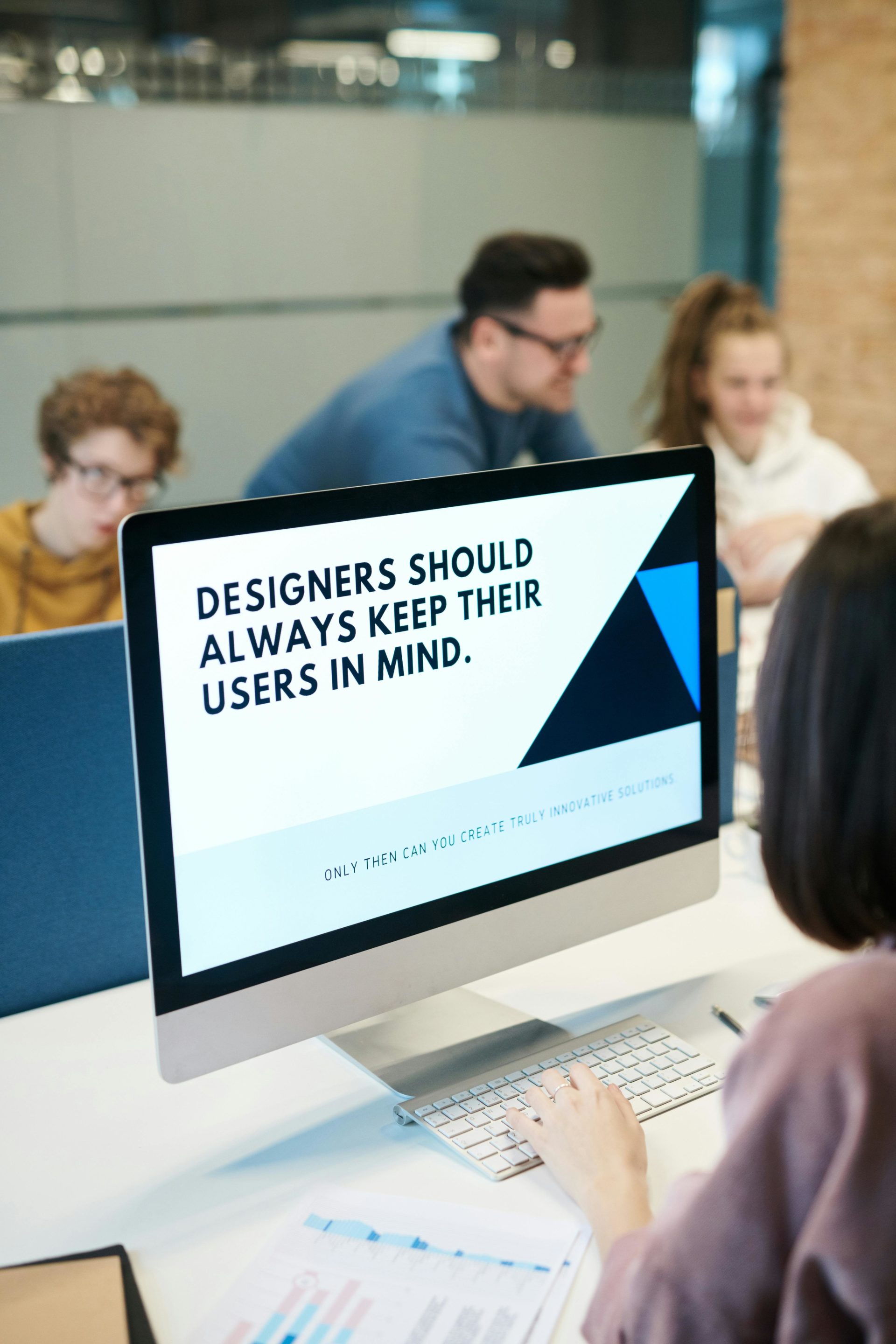 A group of designers attentively discusses around a computer screen displaying a message about user-focused design.