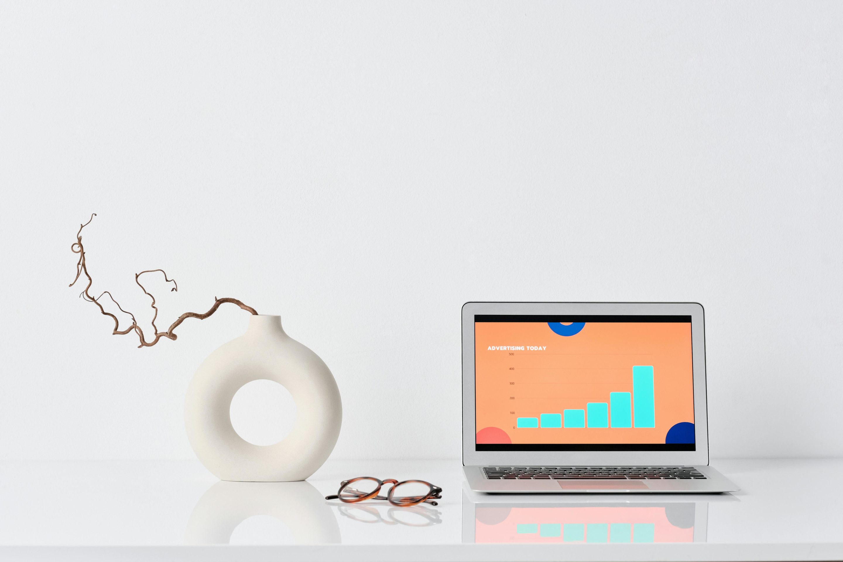 Laptop displaying a bar graph on a white desk beside a modern ceramic vase with dried twigs and a pair of glasses.