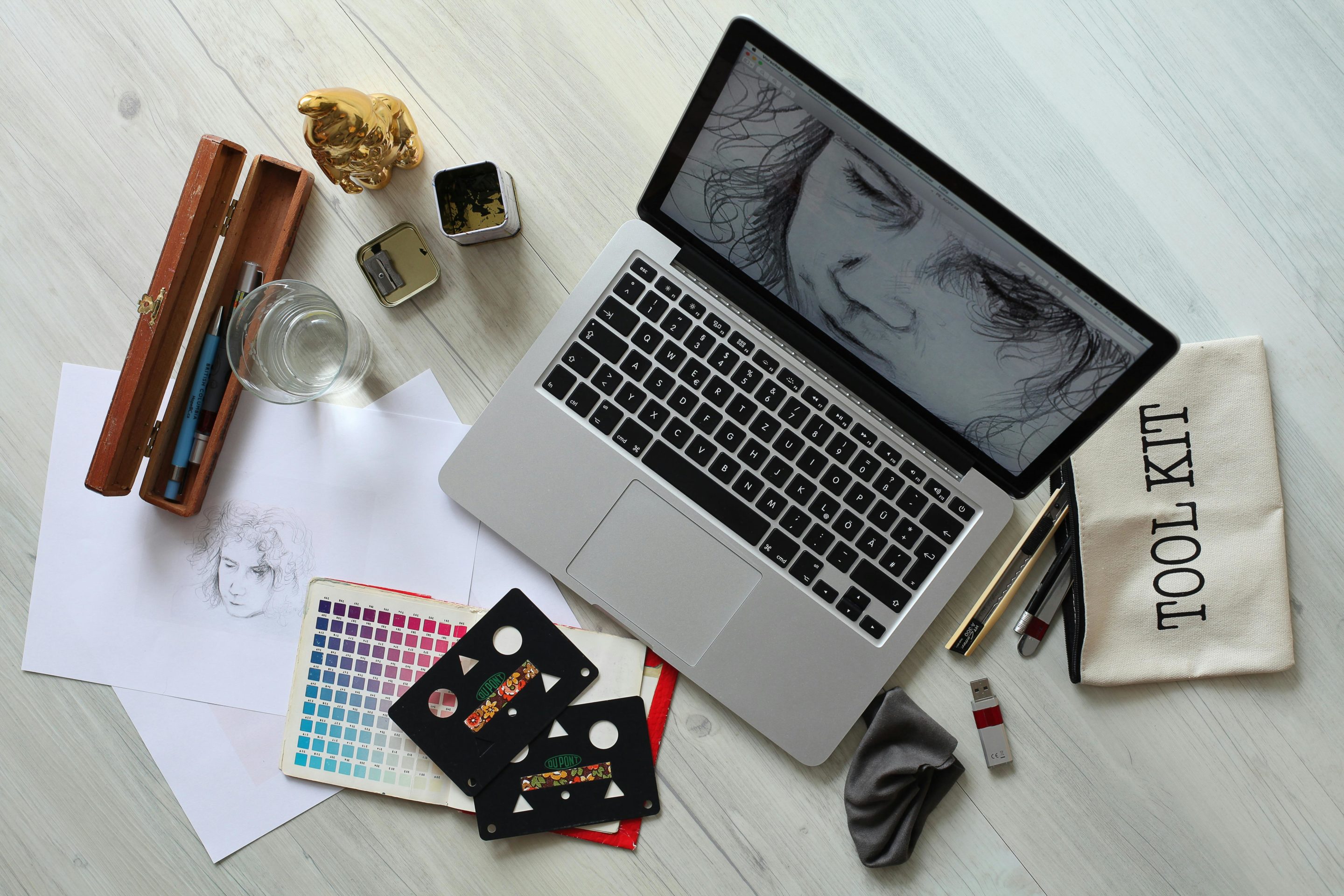 Overhead view of a creative workspace with an open laptop displaying a drawing, sketches, color swatches, and various art tools on a wooden surface.