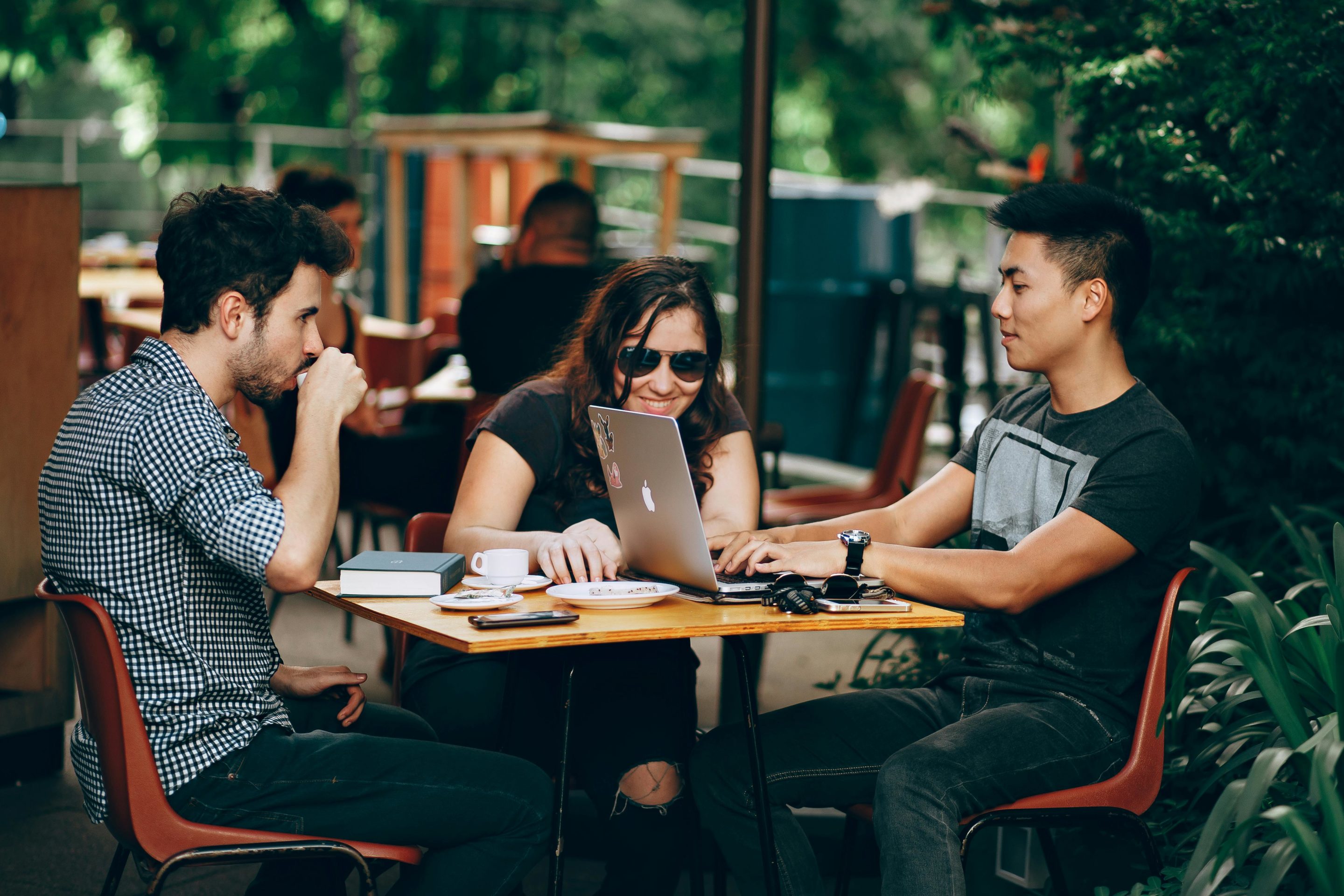 Three young adults, one woman and two men, sit at a café table with laptops and coffee, engaged in a discussion outdoors. Three young adults, one woman and two men, sit at a café table with laptops and coffee, engaged in a discussion outdoors.