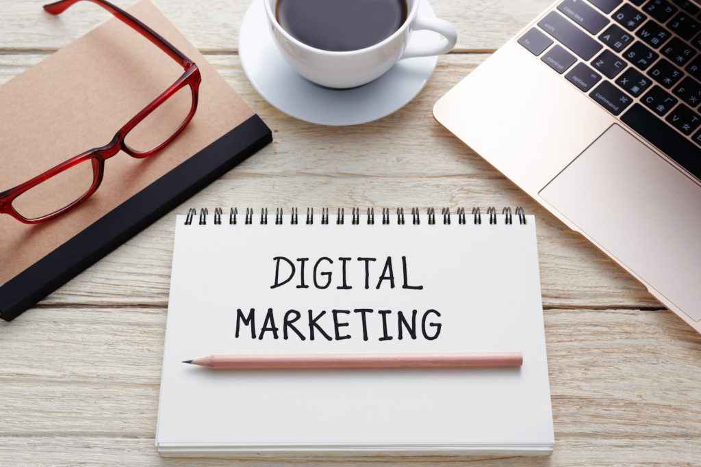 A notebook with "DIGITAL MARKETING" written on it lays on a table in Petaluma CA, accompanied by a pencil, red glasses, a coffee cup, and a laptop.