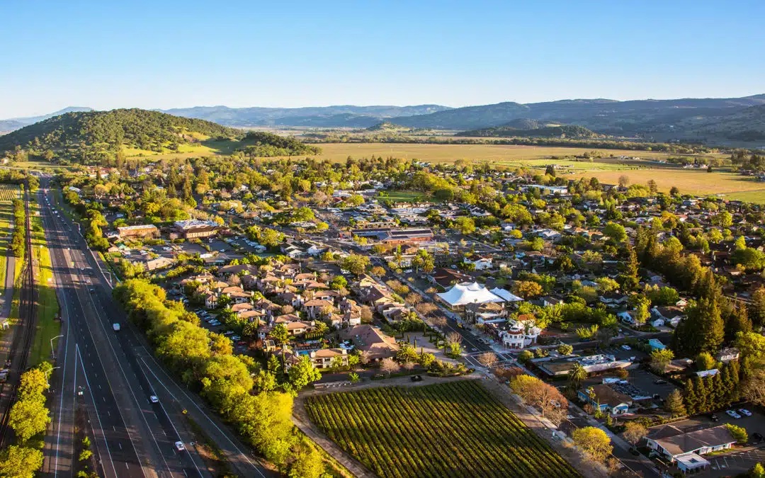 Aerial view of a small town surrounded by green fields and hills, with a highway running alongside residential neighborhoods, trees, and mountains in the background under a clear blue sky.