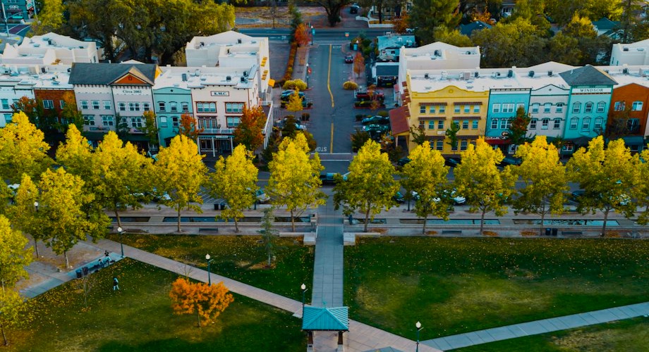 Aerial view of a small town square with green grass, tree-lined walkways, and colorful storefronts in the background during autumn.