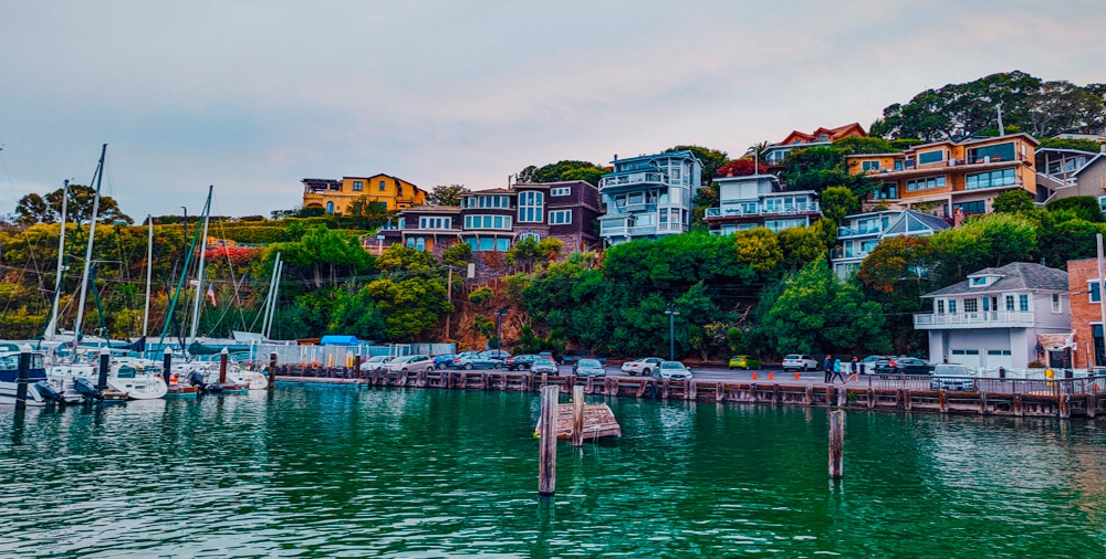 Colorful houses sit on a lush green hillside above a marina with parked boats and cars along the waterfront on a calm, cloudy day.