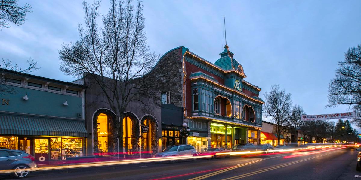 A historic downtown street at dusk features old-fashioned buildings with lit windows, light trails from passing cars, bare trees, and a prominent building with a clock tower.