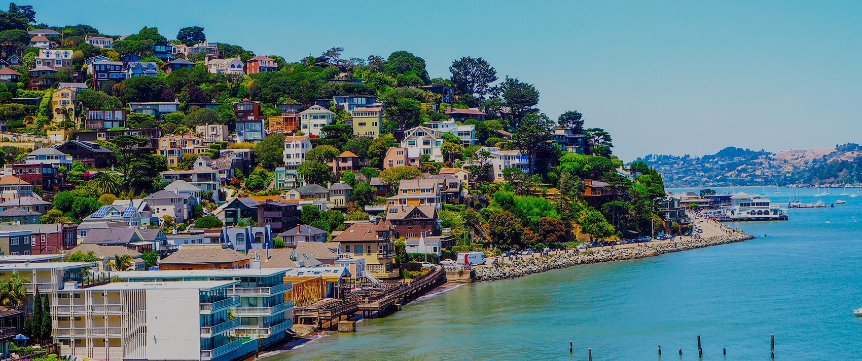 Colorful houses are built on a lush hillside overlooking a calm bay, with docks, boats, and waterfront buildings visible in the foreground and distant hills across the water under a clear blue sky.