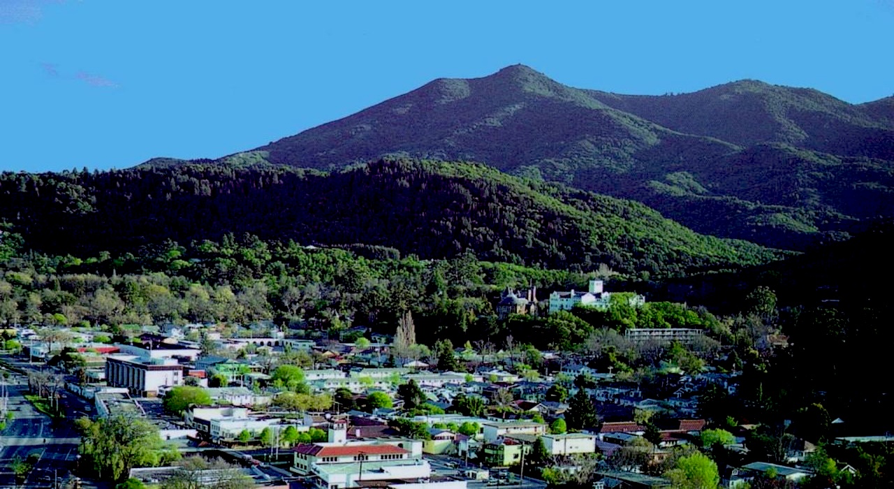 A small town with colorful buildings and trees sits at the base of a forested mountain range under a clear blue sky.