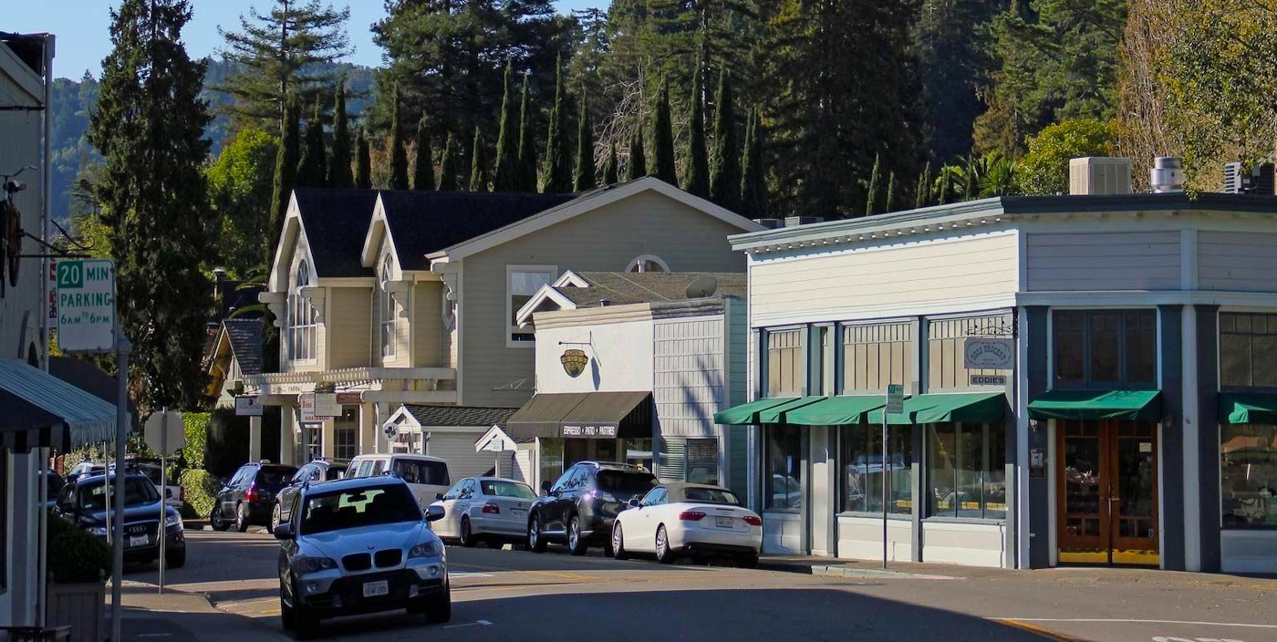 A small town street lined with parked cars, shops, and trees. Buildings have large windows and awnings, and tall evergreen trees are visible in the background under a clear sky.