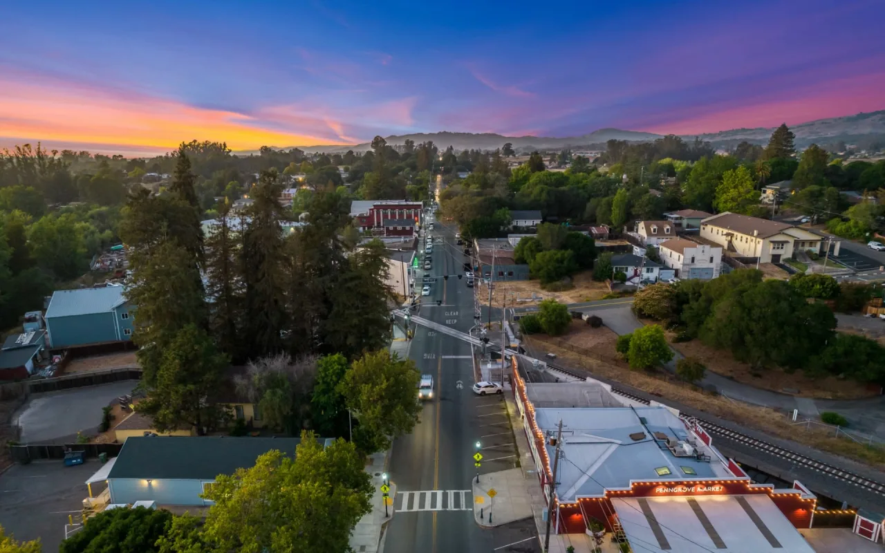 Aerial view of a small town at sunset, featuring a main road lined with buildings and trees, with hills and a colorful sky in the background. Streetlights and cars are visible along the quiet streets.