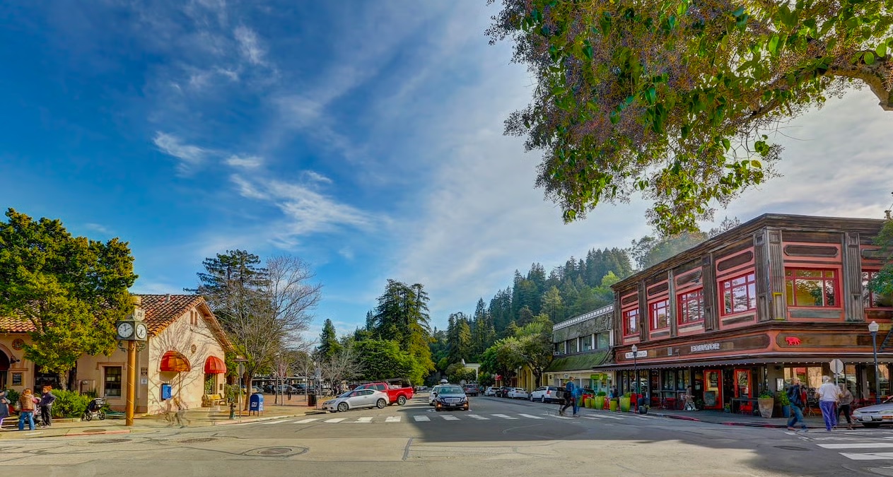 A small-town street scene with shops, trees, parked cars, and people walking. A two-story building with red trim is on the right, and a clock stands near a sidewalk on the left. The sky is partly cloudy.