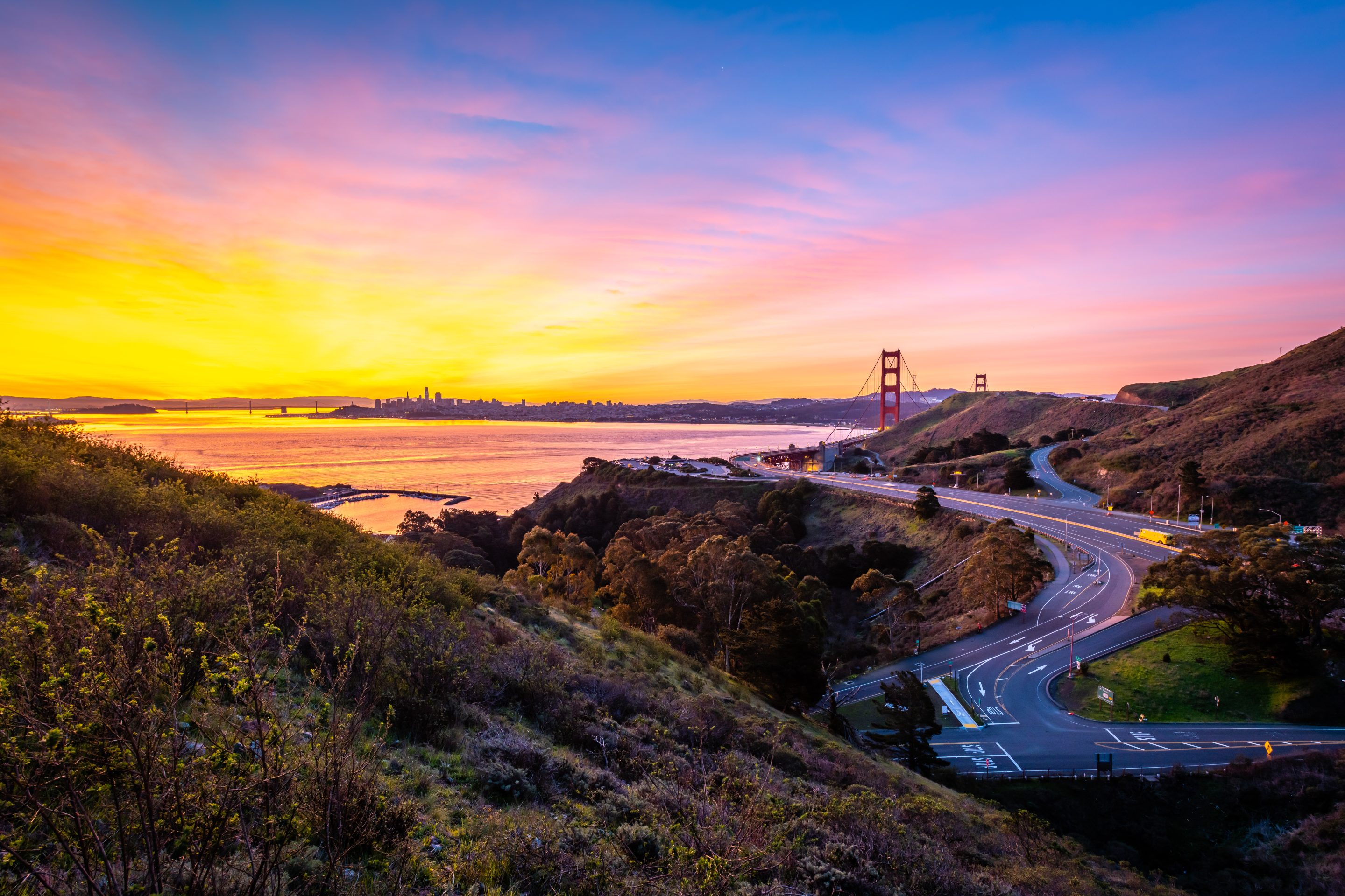 View of the Golden Gate Bridge at sunset from Marin County, with vibrant orange, pink, and purple sky, winding roads in the foreground, and San Francisco skyline across the bay.