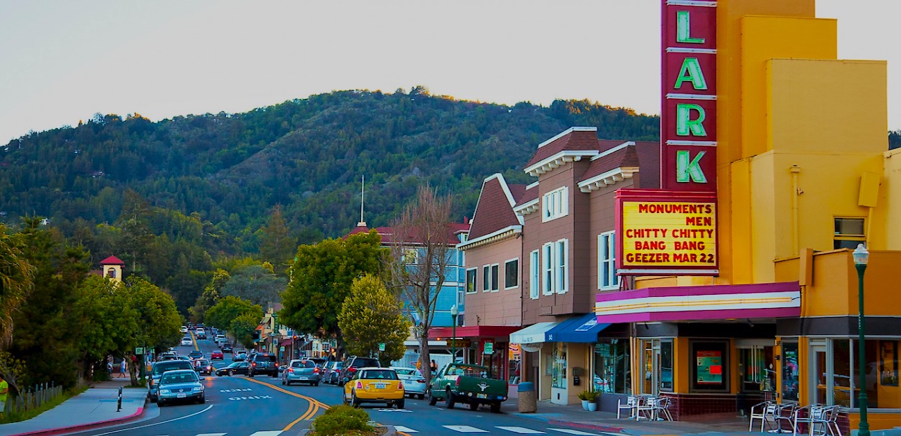 A colorful small-town street with shops, cars parked along the road, and a bright vintage theater sign reading "LARK." Green hills and trees rise in the background under a blue sky.