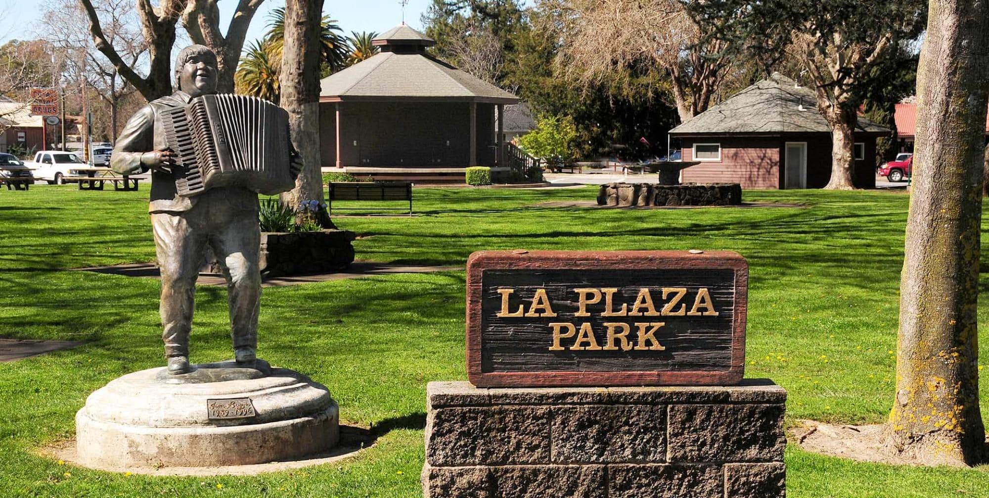 A stone statue of a man playing an accordion stands in a green park near a wooden sign that reads "LA PLAZA PARK." Trees and small buildings are visible in the background on a sunny day.