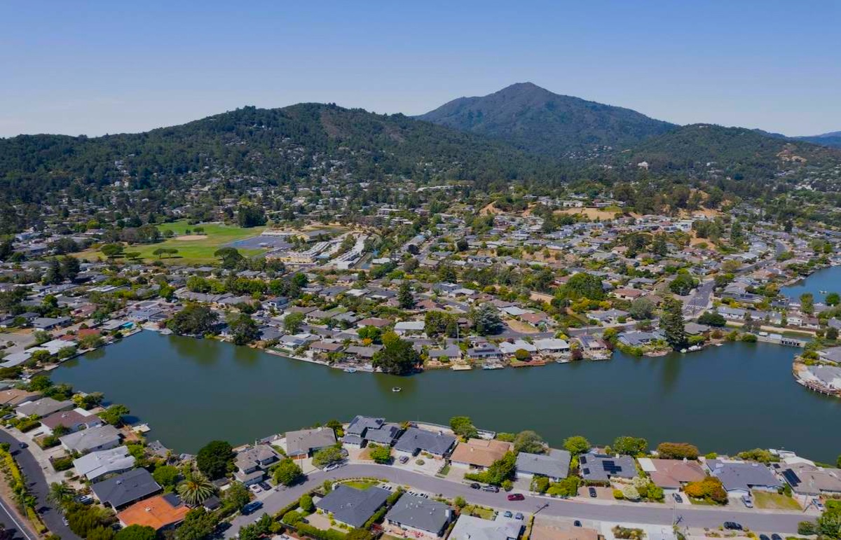 Aerial view of Corte Madera, a suburban neighborhood with houses lining a curved waterfront, surrounded by trees and green hills under a clear blue sky.