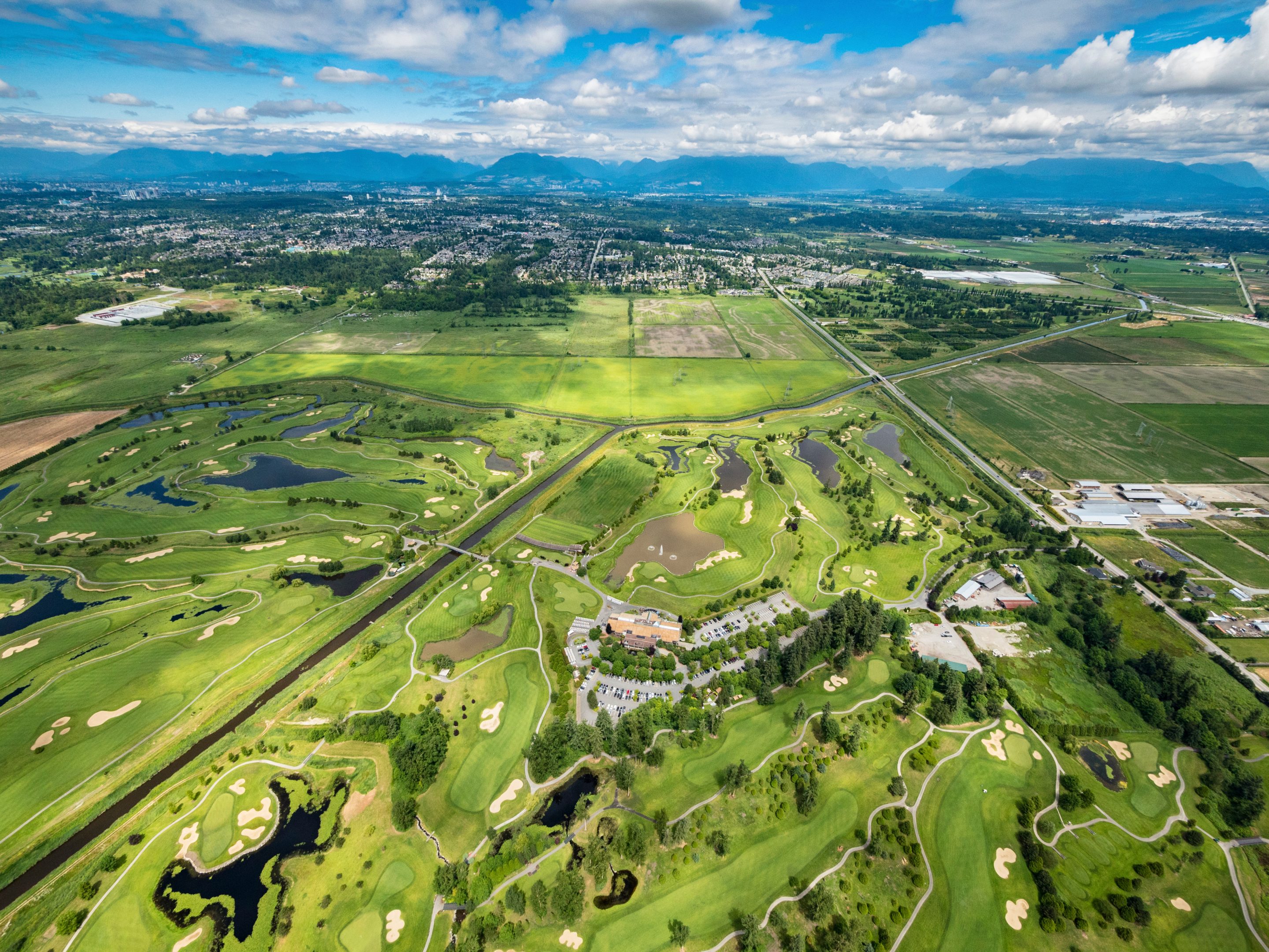 Aerial view of a lush green golf course with sand traps, water features, and a clubhouse, surrounded by fields, roads, and a distant town under a partly cloudy sky.