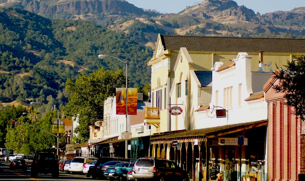 A row of historic buildings with shops and restaurants lines a small town street, with parked cars and tree-covered mountains rising in the background under a clear sky.