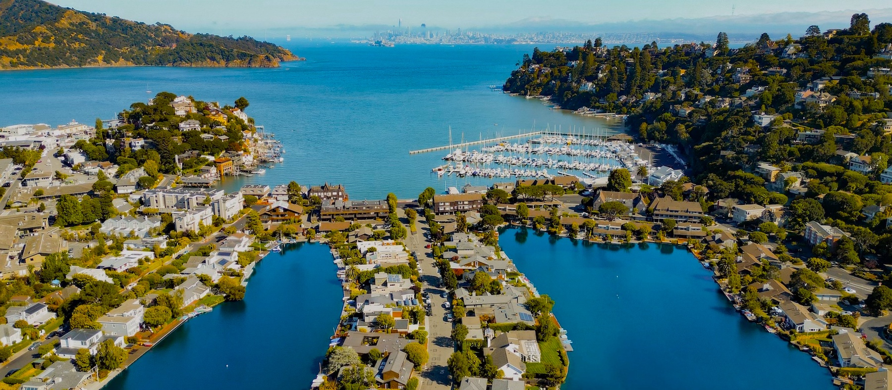 Aerial view of a coastal town with houses along waterways, a marina filled with boats, tree-covered hills, and a city skyline in the distant background across the bay.