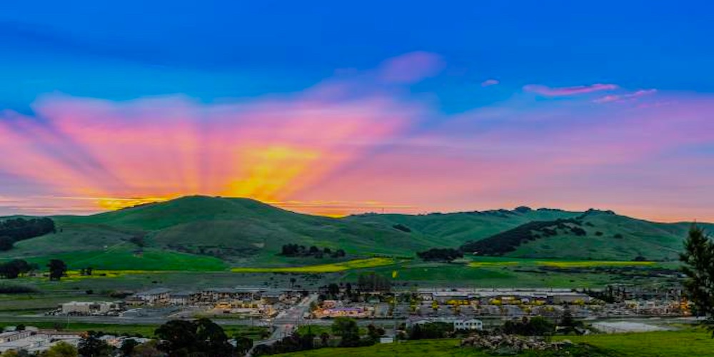 A vibrant sunset casts pink, orange, and yellow rays over green hills near American Canyon, with a small town and roads in the foreground under a clear blue sky.