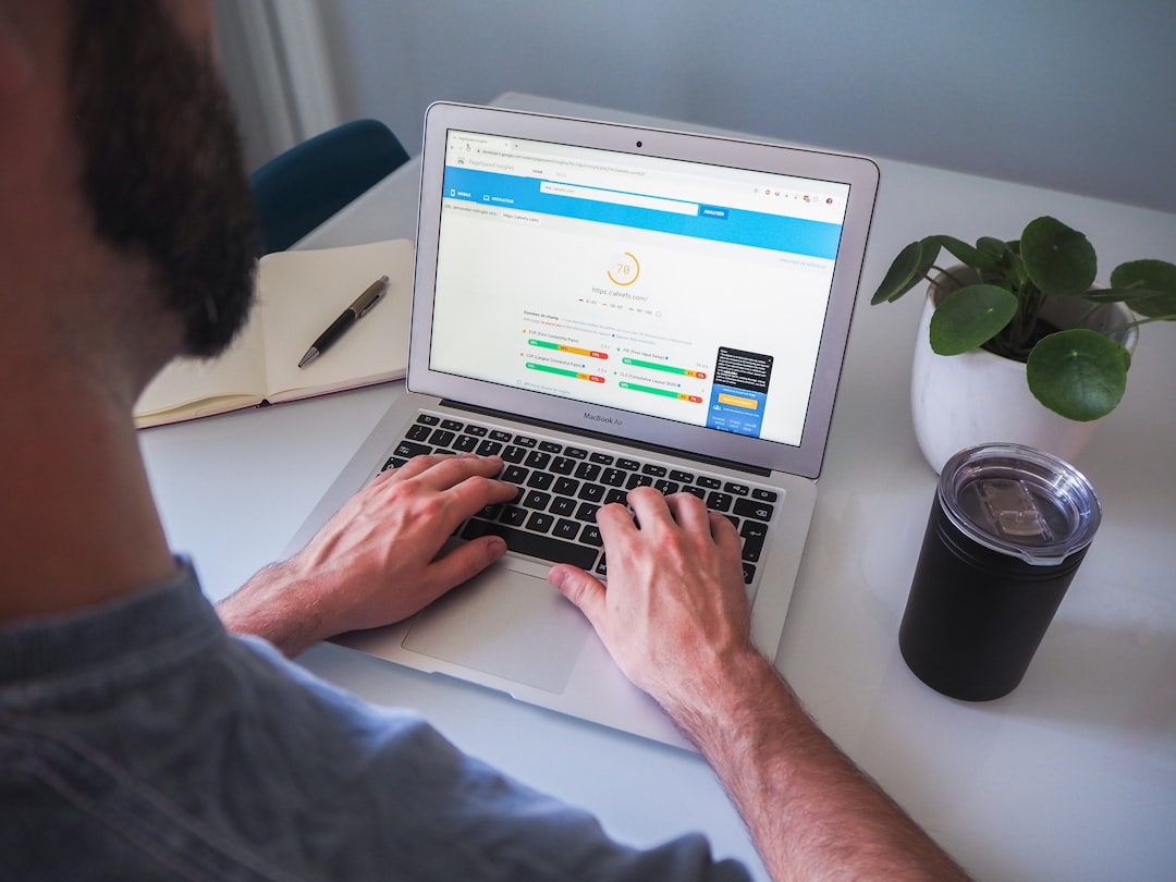 A person types on a laptop displaying a website with SEO performance metrics. A notebook, pen, plant, and insulated tumbler sit on the white desk next to the laptop.